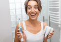 Smiling woman holding an electric toothbrush and a large tooth model in a bathroom.