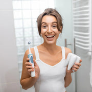 Smiling woman holding an electric toothbrush and a large tooth model in a bathroom.